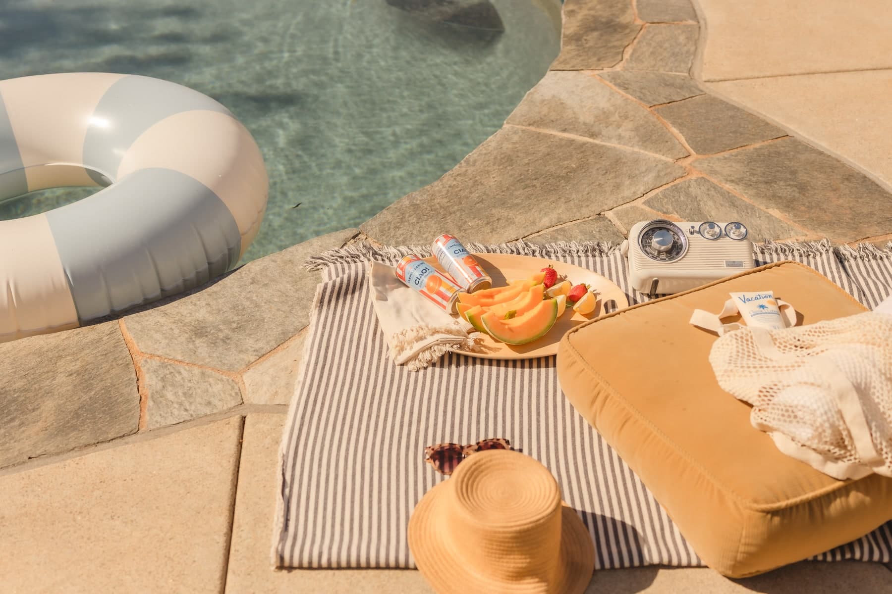 Poolside picnic with fruit, towels, a sun hat, and an inflatable ring beside the water